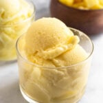 A glass cup with scoops of pineapple dole whip in it. Behind it is another glass cup filled and a bowl of frozen pineapple.
