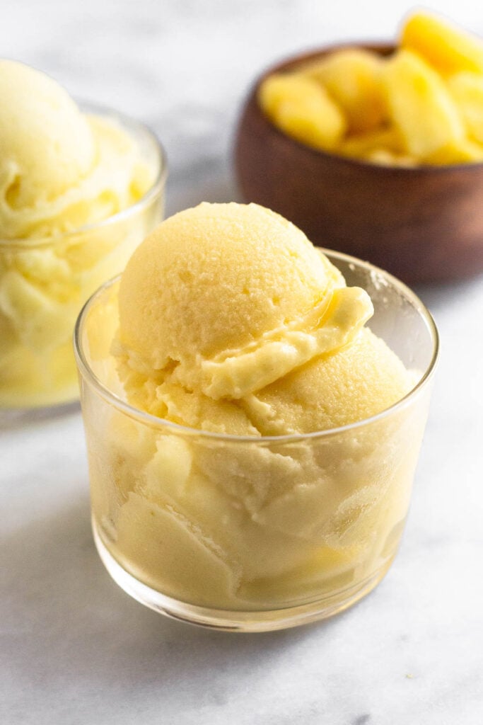 A glass cup with scoops of pineapple dole whip in it. Behind it is another glass cup filled and a bowl of frozen pineapple.