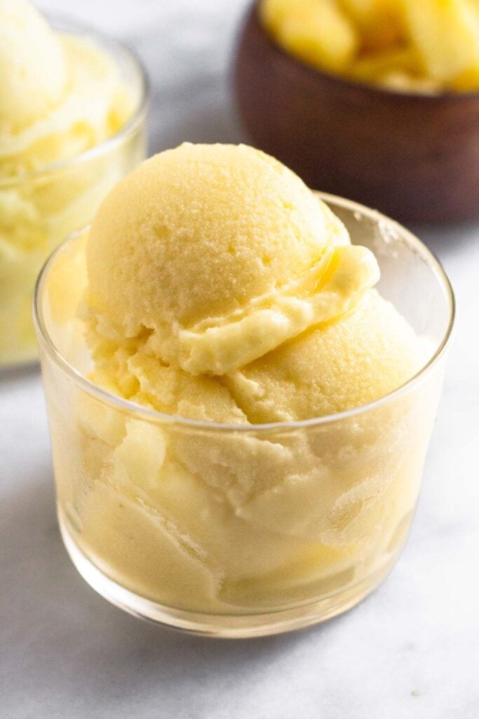 Scoops of pineapple dole whip in a small glass cup. Behind it is another cup and a bowl of frozen pineapple.