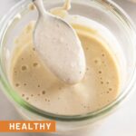 Healthy caesar dressing in a jar with a spoon coming out of it. In front of the jar is half a juiced lemon and behind it is a bowl of salad.