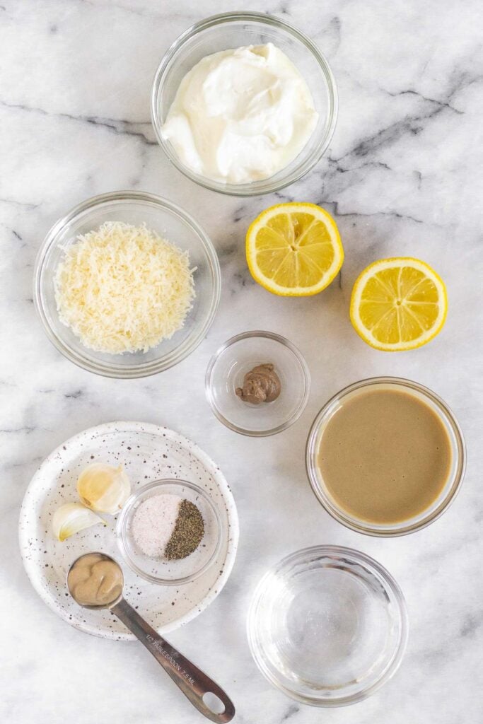 White marble counter with a bowl of greek yogurt, a lemon cut in half, a bowl of tahini, a bowl water, a plate with a teaspoon of mustard, 2 garlic cloves, and a bowl of salt and pepper, a bowl of anchovy paste, and a bowl of grated parmesan cheese.
