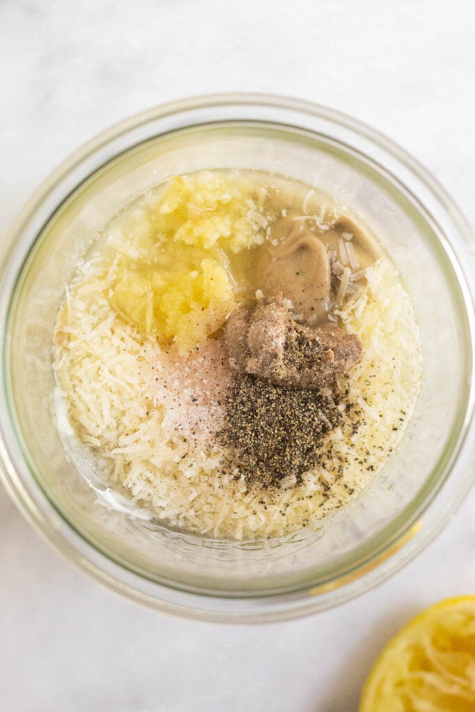 Overhead shot of a large jar with grated parmesan cheese, minced garlic, dijon mustard, anchovy paste, salt, and pepper in it. Next to is it a half a juiced lemon.