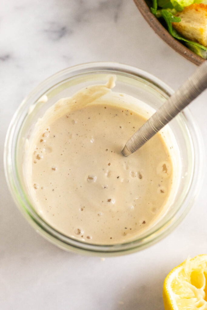 A jar of greek yogurt caesar dressing with a spoon in it. Next to it is a bowl of salad and half a juiced lemon.