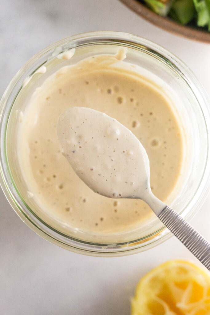 Healthy Caesar salad dressing in a jar with a spoon overtop holding some. Off to the side is a bowl of salad and half a juiced lemon.