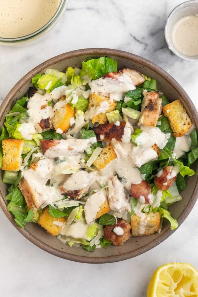 A bowl of salad with chopped romaine, grilled chicken, croutons, parmesan cheese, and yogurt caesar dressing on top. Around the bowl is a jar of dressing, a small ramekin of dressing, and half a lemon wedge.