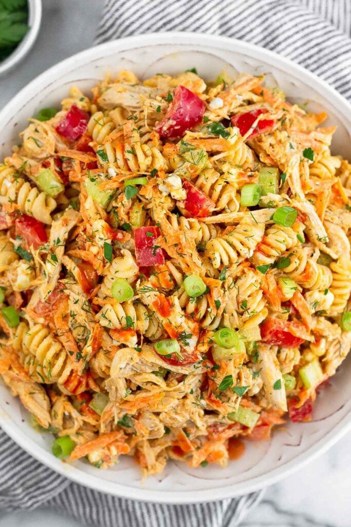Buffalo chicken protein pasta salad with veggies in a large bowl. The bowl is sitting on a striped towel with a bowl of herbs around it.