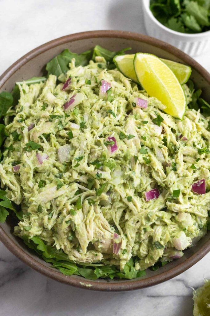 Avocado chicken salad on top of some fresh arugula in a bowl. It is garnished with 2 lime wedges. Next to the bowl is half a juiced lime and a bowl of cilantro.