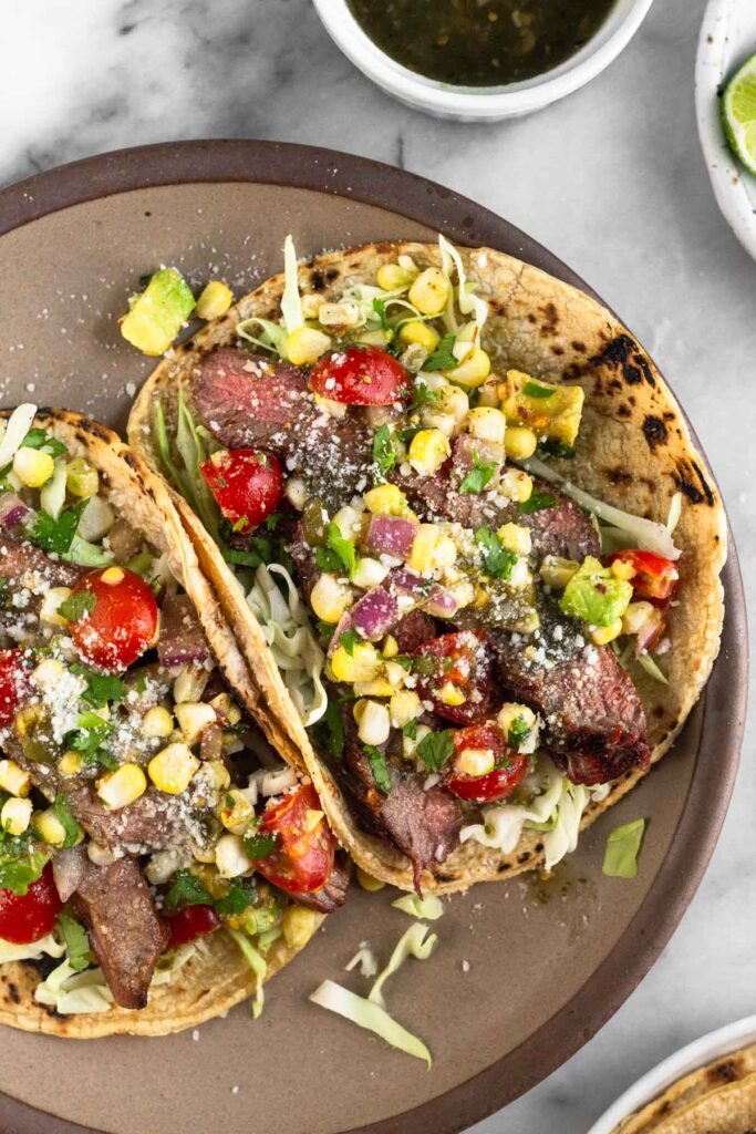Overhead shot of two steak tacos on a brown plate. They are stuffed with cabbage, corn, tomatoes, avocado, and cilantro. Next to the plate is a bowl of salsa, a plate of limes, and plate of tortillas.