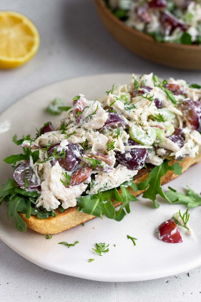 Healthy chicken salad on top of some arugula on a piece of toast on a plate. Behind it is half a lemon and a bowl of more chicken salad.
