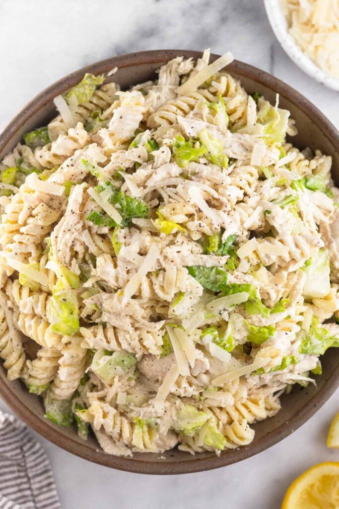 Chicken caesar pasta salad in a large brown bowl. Next to the bowl is half a juiced lemon, a bowl of grated parmesan cheese, and a kitchen towel.