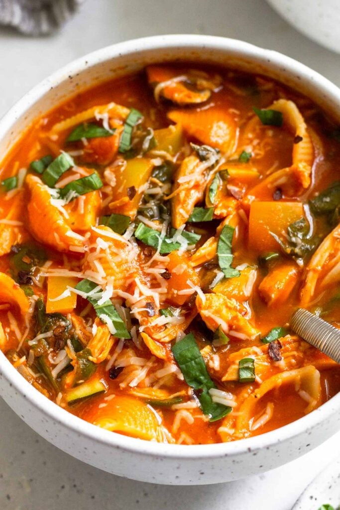 A bowl of tomato soup with shredded chicken and veggies topped with basil and parmesan cheese. There is a spoon in the bowl.