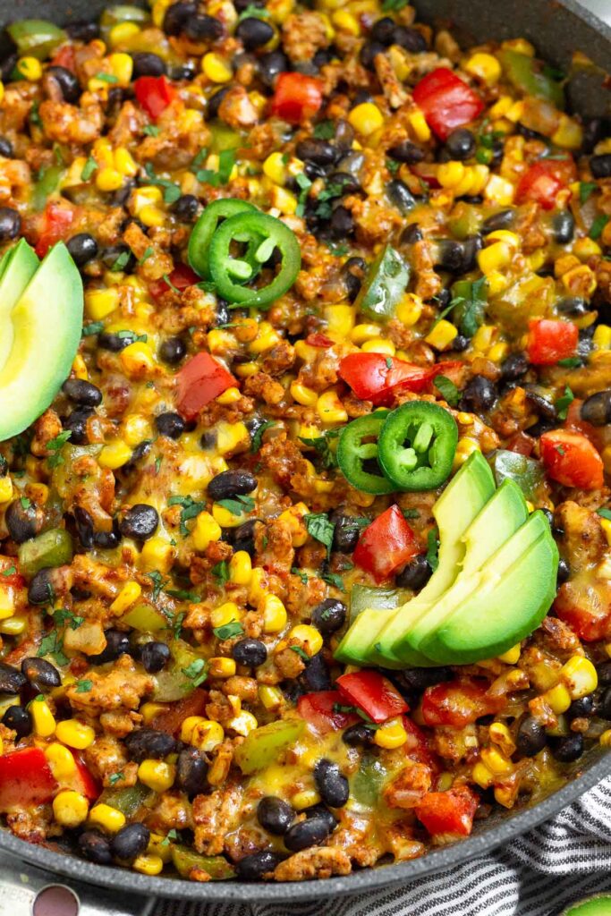 Close up of a skillet filled with chicken, corn, veggies, black beans, and melted cheese. It is topped with cilantro, jalapeños, and avocado.