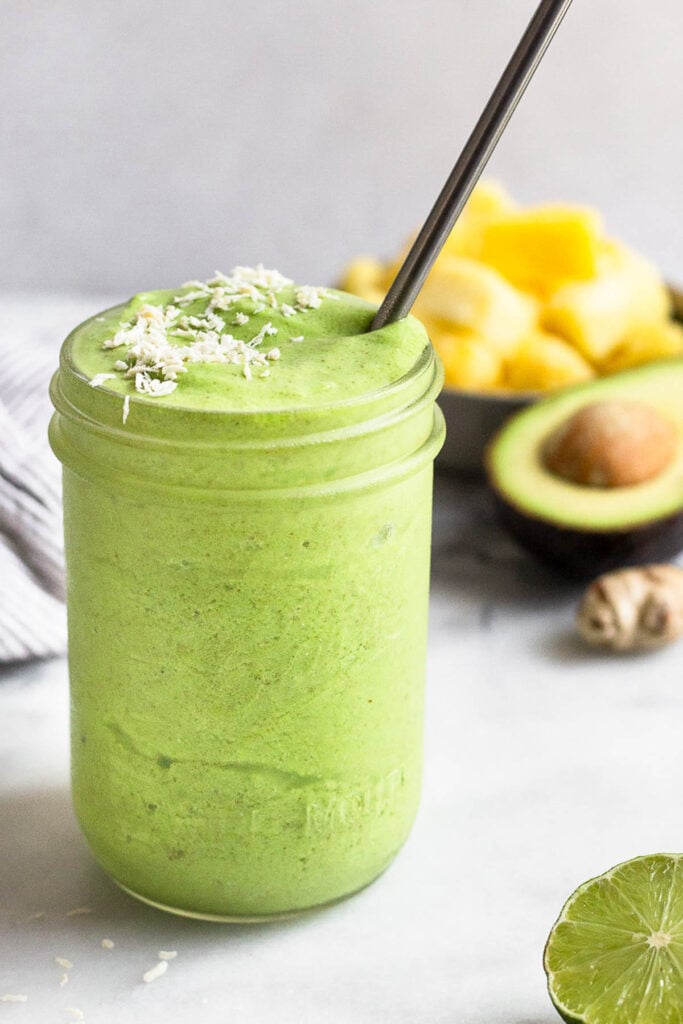 Green protein smoothie in a glass jar topped with shredded coconut and a straw. Around it is half a lime wedge, half an avocado, fresh ginger, and a bowl of frozen pineapple.