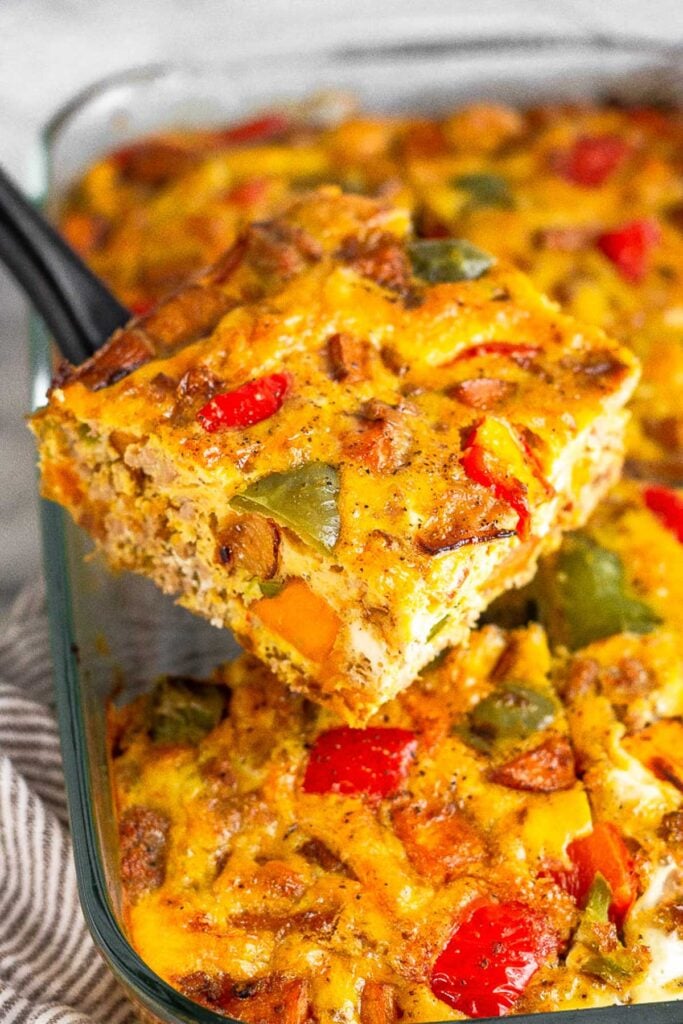 Healthy breakfast casserole with veggies, sweet potatoes, and sausage in a pan. One piece is being held up by a spatula above the rest.