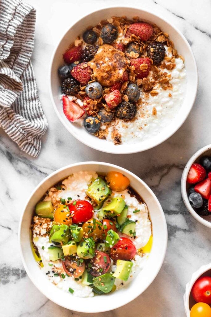 Overhead shot of two cottage cheese bowls. One has berries, peanut butter, and granola and the other has tomatoes, cucumber, and avocado. Next to them is a kitchen towel, a bowl of berries, and a bowl of tomatoes.
