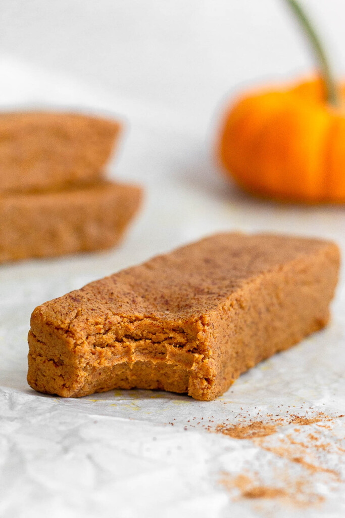 A healthy pumpkin protein bar with a bite taken out of it. Behind it is two bars stacked on top with each other and a small pumpkin.