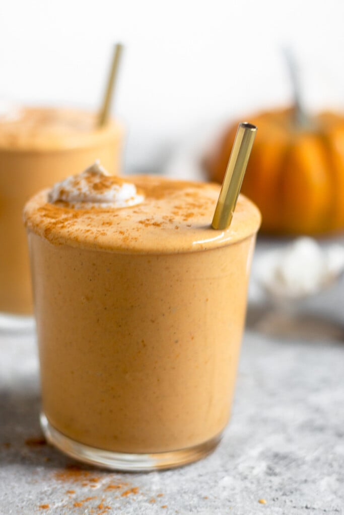 Pumpkin milkshake in a glass cup topped with whipped cream and cinnamon with a straw sticking out of it. Behind it is another full glass and a pumpkin.
