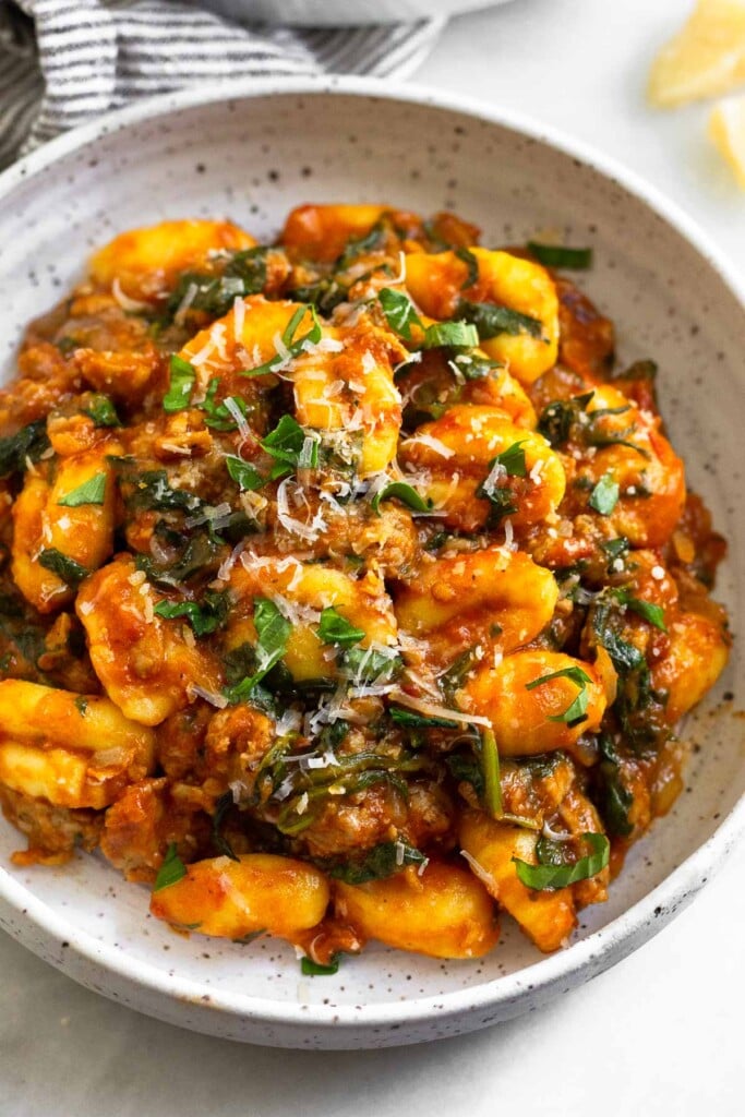White plate with gnocchi & Italian sausage mixed with pasta sauce. It is garnished with basil and parmesan cheese.