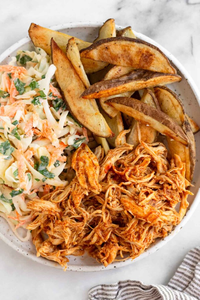 A white plate of pulled BBQ chicken, coleslaw, and potato wedges. Next to the plate is a striped kitchen towel.