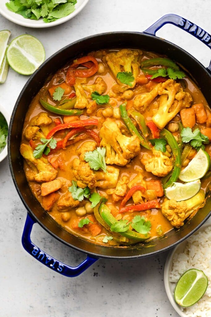 Large dutch oven filled with vegan cauliflower chickpea curry garnished with cilantro and lime wedges. Next to it is lime wedges and a bowl of white rice.