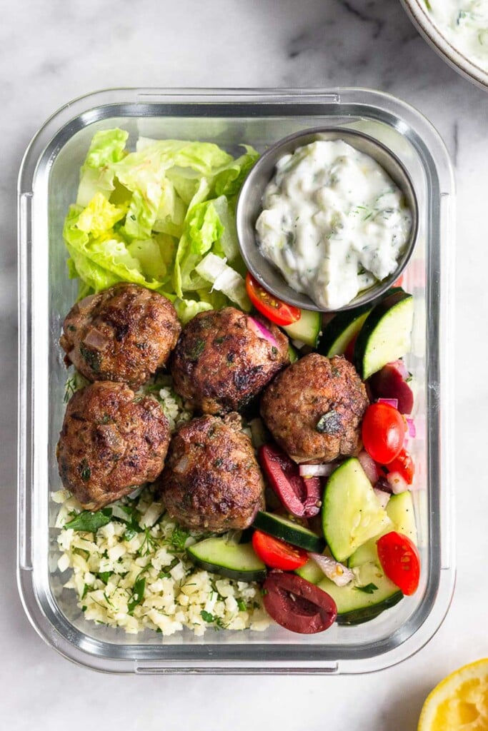 High protein lunch recipe with romaine lettuce, herby rice, a tomato, cucumber, and olive salad, greek lamb meatballs, and a small ramekin of tzatziki sauce all in a glass meal prep container.