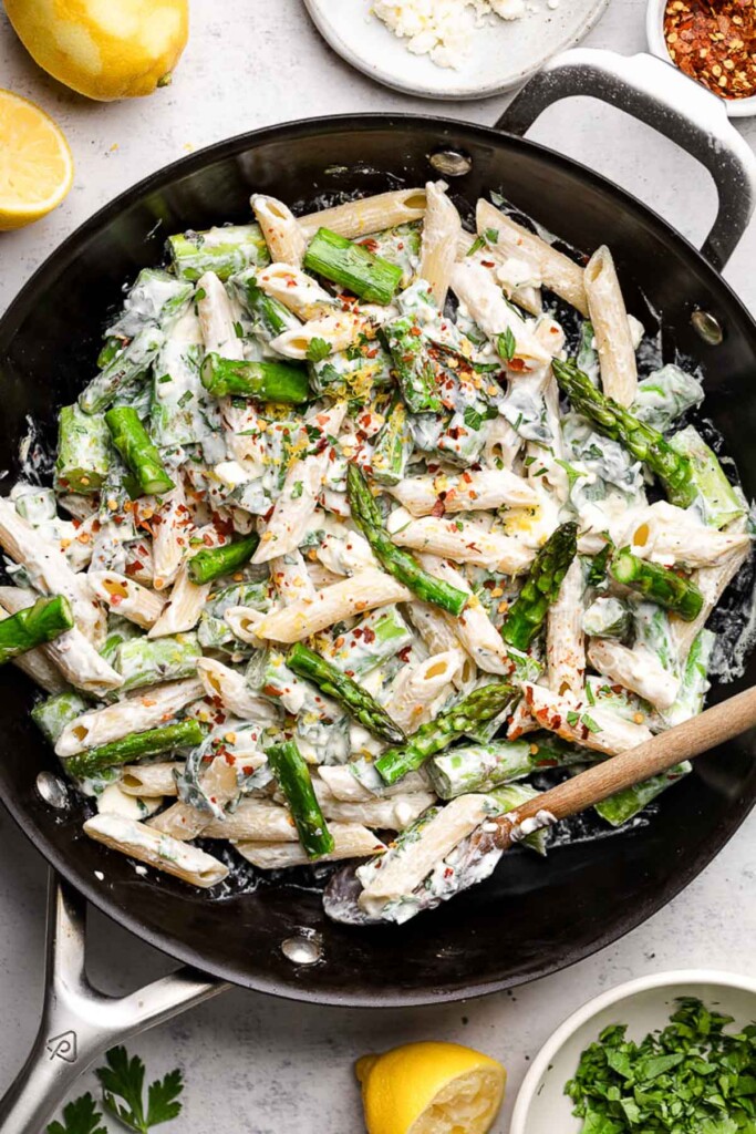 Pan of lemon asparagus pasta with creamy feta sauce topped with red pepper flakes. Around the pan is some lemons, herbs, and spices.