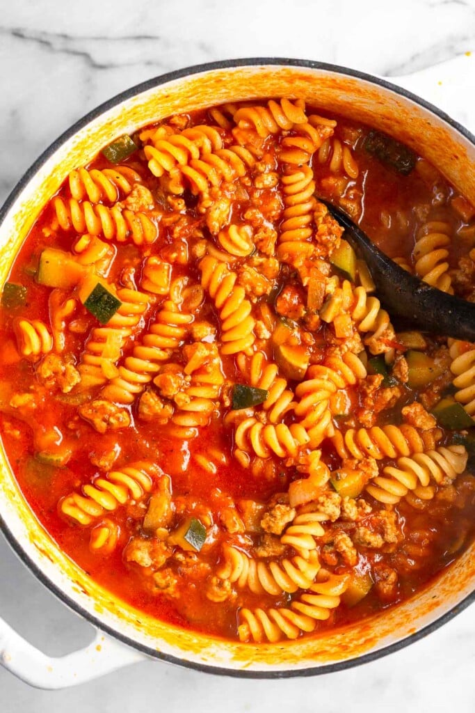 One pot pasta with meat and veggies in a white dutch oven. A spoon is in the pot.