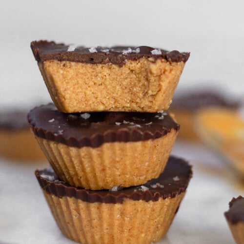 Three protein peanut butter cups stacked on top of each other on a white marble counter. The top one has a bite taken out of it. Around them are more PB cups.