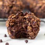 Chocolate oatmeal muffin with a bite taken out of it with some mini chocolate chips around it. Behind it is a rack of more muffins.