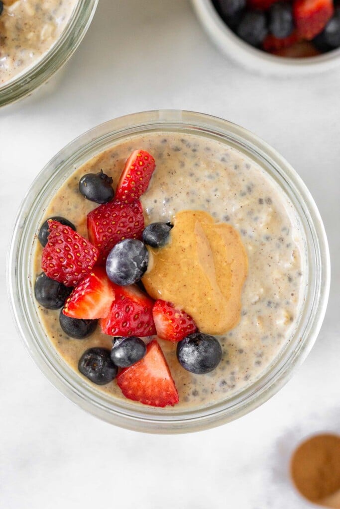 Overhead shot of a jar of overnight protein oats topped with diced strawberries, blueberries, and peanut butter. Next to it is other jar of overnight oats, a teaspoon of cinnamon, and a ramekin of berries.