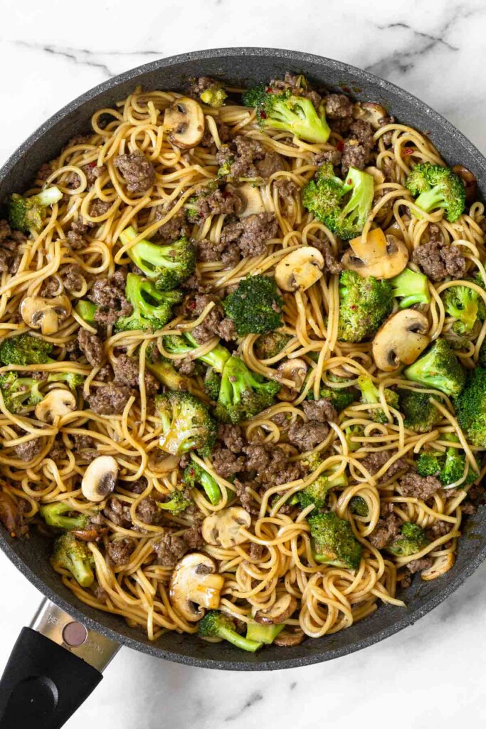 Ground beef and ramen noodles with broccoli and mushrooms tossed in sauce in a large skillet.