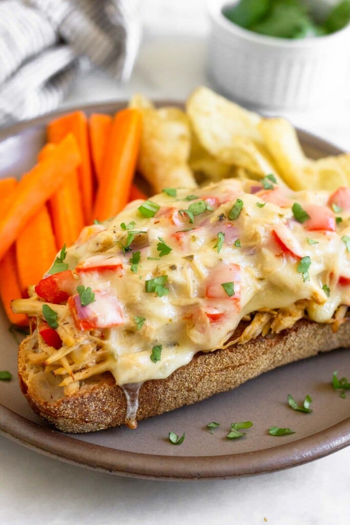 Chicken salad melt garnished with cilantro on a brown plate with sliced carrots and potato chips. Behind the plate is a striped kitchen towel and a bowl of cilantro.