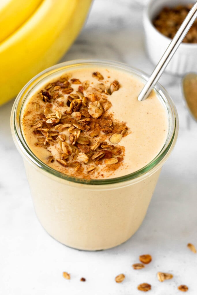 Peanut butter banana protein smoothie in a glass jar topped with granola and cinnamon with a straw in it. Around the jar are some bananas and a bowl of granola.