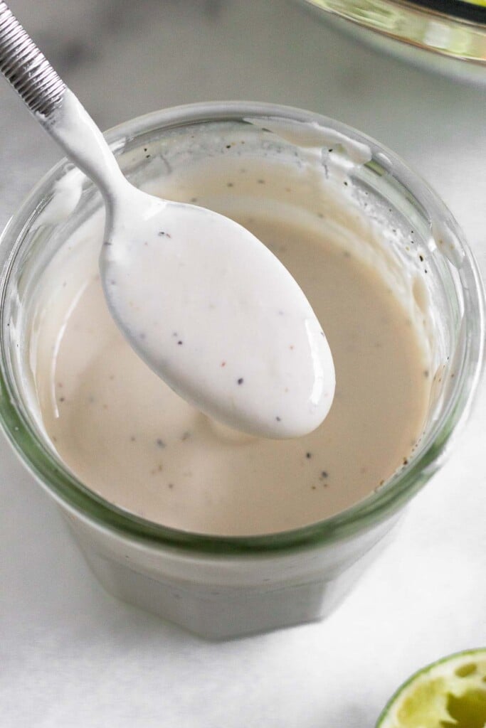 Healthy coleslaw dressing in a jar with a spoon full of dressing overtop of it. In front of the jar is half a juiced lime.