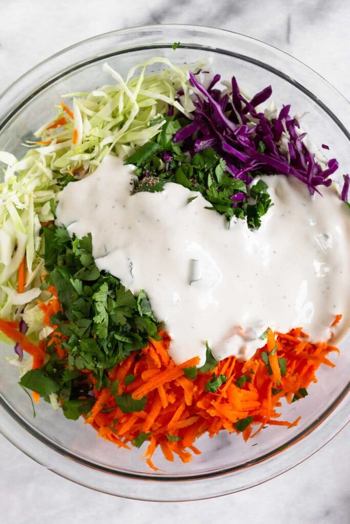 Large glass bowl filled with shredded green and red cabbage, shredded carrots, and cilantro with a yogurt dressing poured overtop before it is mixed together.
