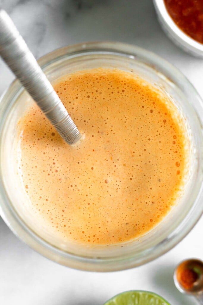 A jar fill of salsa dressing with a spoon in it. Around the jar is a dish of salsa, a teaspoon with spices in it, and half a lime.