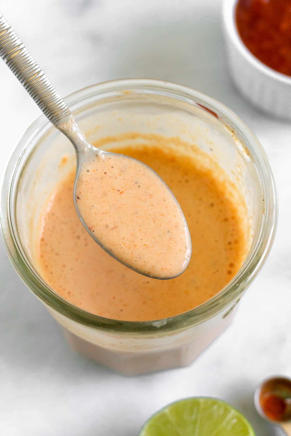 Creamy salsa dressing in a jar with a spoon full of dressing overtop of it. In front of the jar is half a lime and a teaspoon with spices in it. Behind the jar is a small dish of salsa.