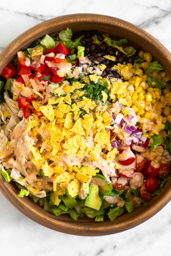 Large wooden salad bowl with chopped romaine, shredded chicken, diced peppers, black beans, corn, cherry tomatoes, red onion, avocado, and cilantro. It is topped with salsa dressing and crushed tortilla chips.