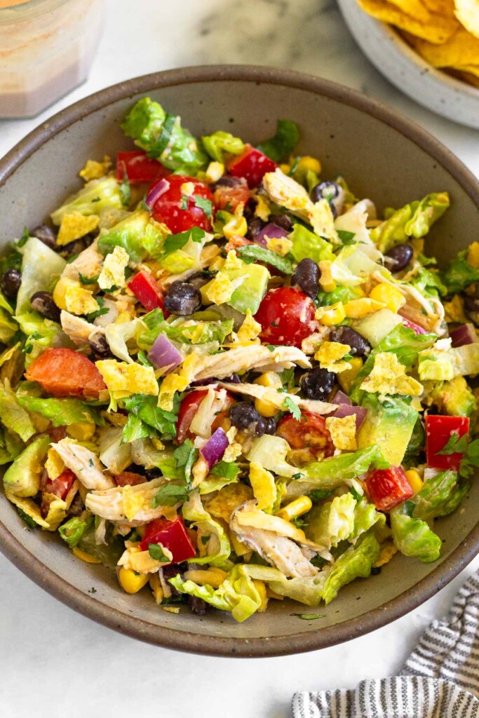 Chopped chicken taco salad with lettuce, tomato, shredded chicken, peppers, avocado, black beans, corn, and chips in a brown bowl. Around to the bowl is a kitchen towel, a bowl of tortilla chips, and a jar of dressing.