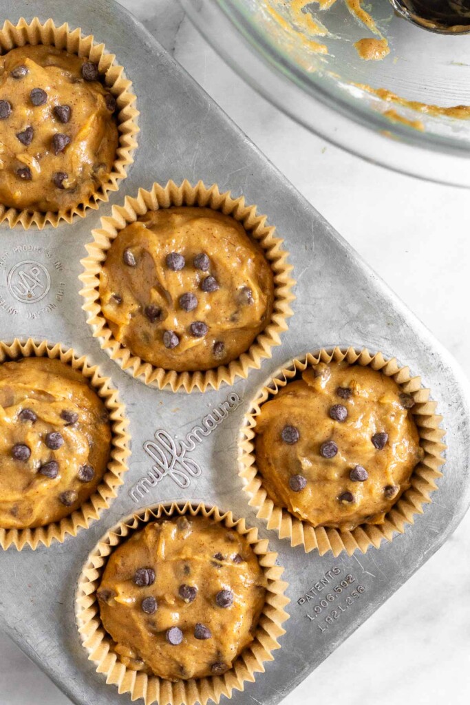 Muffin tin filled with uncooked muffin batter with chocolate chips in it. Off to the side is a empty large bowl with a little batter in it.
