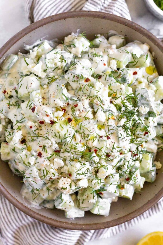 Cucumber yogurt salad garnished with fresh dill, olive oil, and red pepper flakes in a brown bowl. The bowel is still on a striped towel and next to it is half a lemon and a small dish of fresh dill.