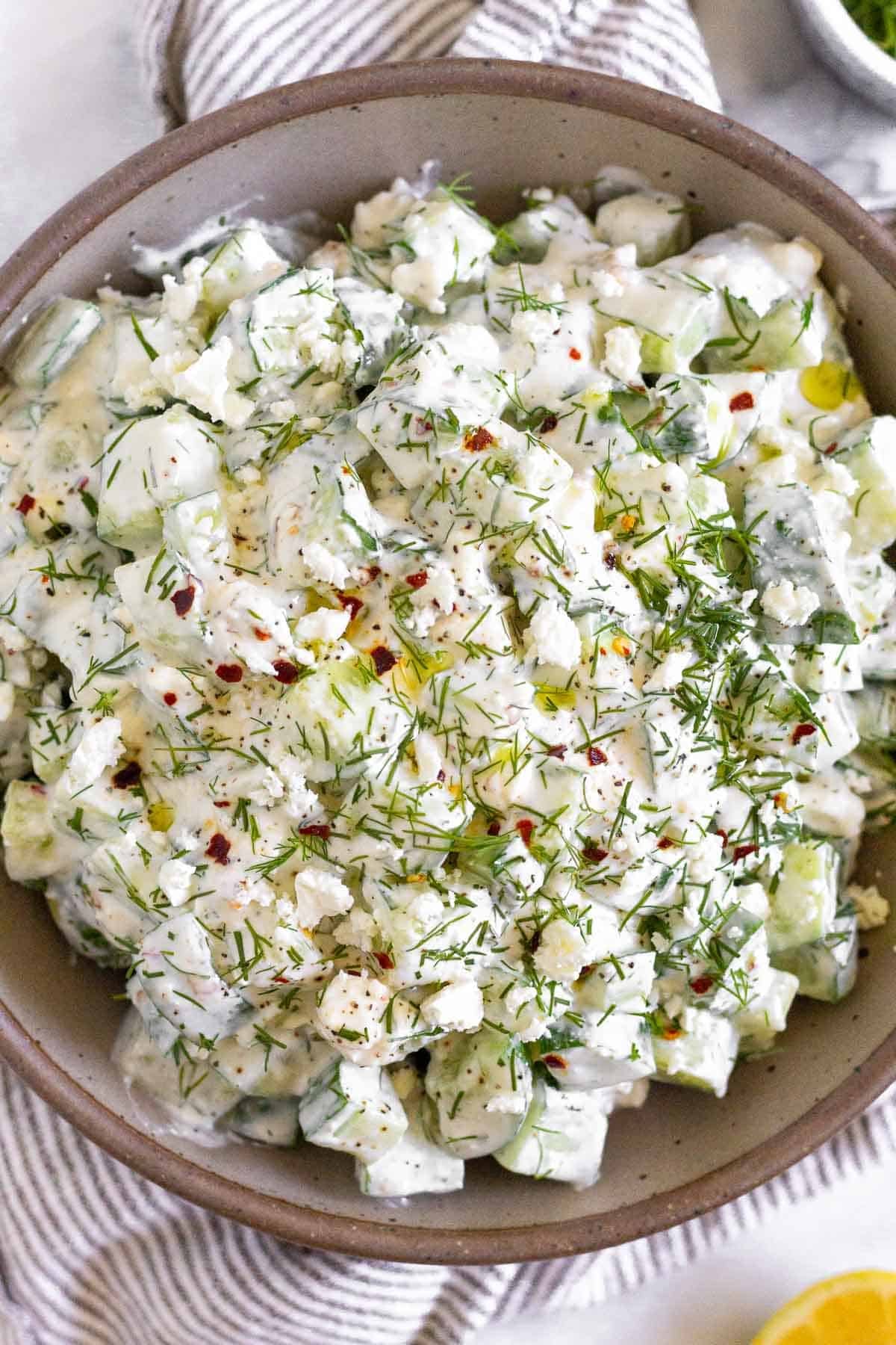 Cucumber yogurt salad garnished with fresh dill, olive oil, and red pepper flakes in a brown bowl. The bowel is still on a striped towel and next to it is half a lemon and a small dish of fresh dill.