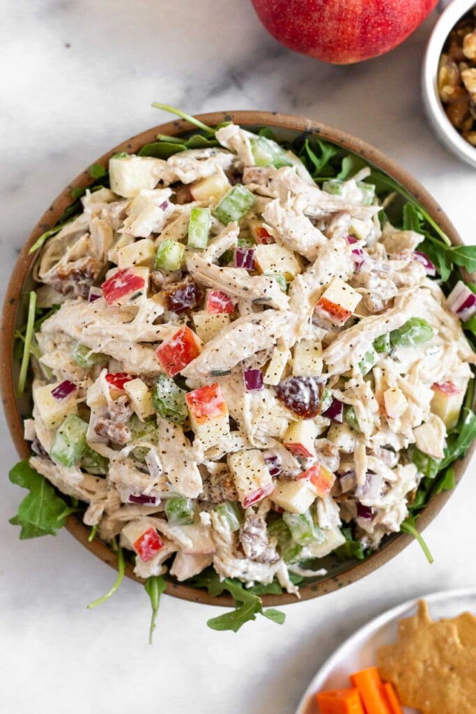 Chicken salad with apples and walnuts in a bowl with arugula underneath it. Around the bowl is a plate of crackers and carrots, a small dish of walnuts, and a red apple.