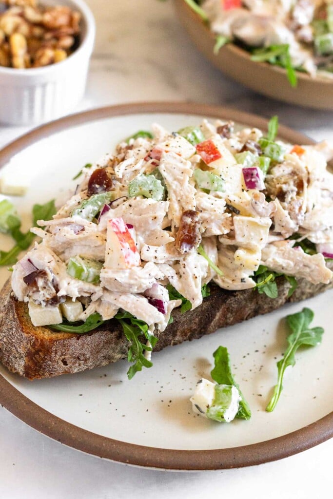 Apple chicken salad with walnuts and celery on top of some arugula on a piece of toast on a plate. Behind it is a small ramekin of walnuts and a bowl of chicken salad.