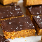 Peanut butter crunch bars topped with chocolate and flaky sea salt lined up on a piece of white parchment paper.