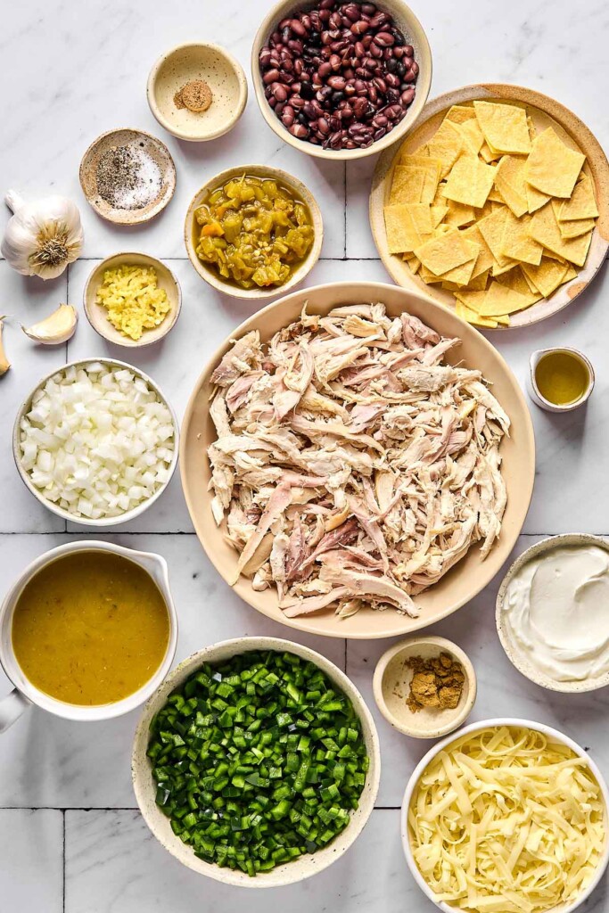 White tile countertop with a bowl of black beans, a bowl of chopped corn tortillas, a small dish of olive oil, a large bowl of shredded chicken, a bowl of greek yogurt, a bowl of spices, a bowl of shredded cheese, a bowl of diced poblano peppers, a bowl of green enchilada sauce, a bowl of diced onions, a bowl of minced garlic, a whole garlic head with a garlic clove next to it, a bowl of diced green chiles, a bowl of salt and pepper, and and bowl of spices.