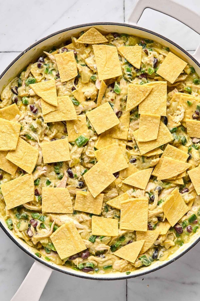 Large while skillet filled with a mixture of shredded chicken, veggies, and black beans in a creamy green sauce with pieces of chopped corn tortillas overtop of it.