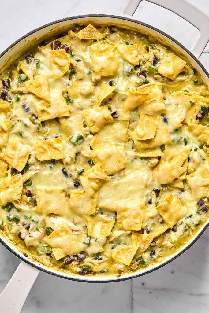 Melted cheese covering chopped corn tortillas overtop a mixture of chicken, beans, and veggies tossed in a creamy green sauce in a large white skillet.