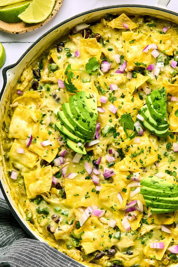 Skillet enchiladas with shredded chicken, beans, veggies, and pieces of corn tortillas covered with melted cheese in a large white skillet. It is garnished with sliced avocado, diced red onions, and cilantro. Next to the pan is a plate of lime wedges.