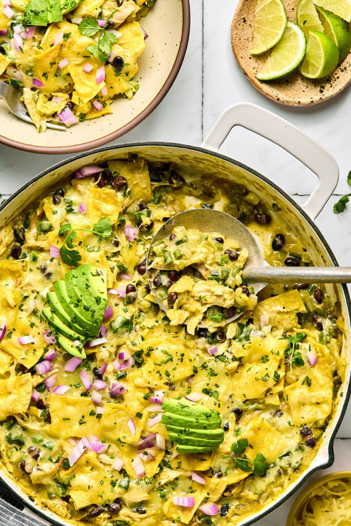 Chicken enchilada skillet with beans and veggies covered in melted cheese in a large white pan. There is a large spoon in the pan with a scoop out of it. It is garnished with sliced avocado, diced red onion, and cilantro. Next to it is a serving in a bowl and a plate of lime wedges.